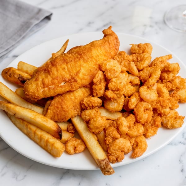 Golden-brown fried fish, crispy shrimp, and french fries on a white plate on a marble table.