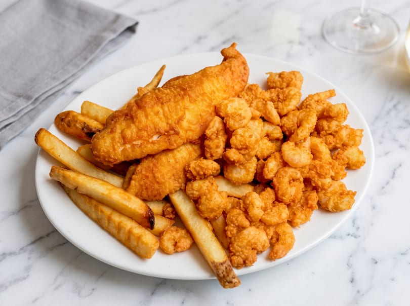 Golden-brown fried fish, crispy shrimp, and french fries on a white plate on a marble table.
